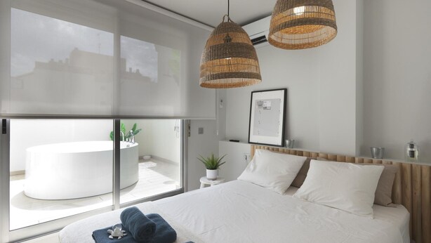 Un dormitorio moderno con una cama grande, dos lámparas colgantes de mimbre, un cabecero blanco, una mesita de noche pequeña con una planta y vistas a un paisaje urbano a través de ventanas de piso a techo.