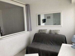 Um quarto pequeno e reservado, com uma cama de solteiro, janela com cortinas, televisão na parede e um espelho.