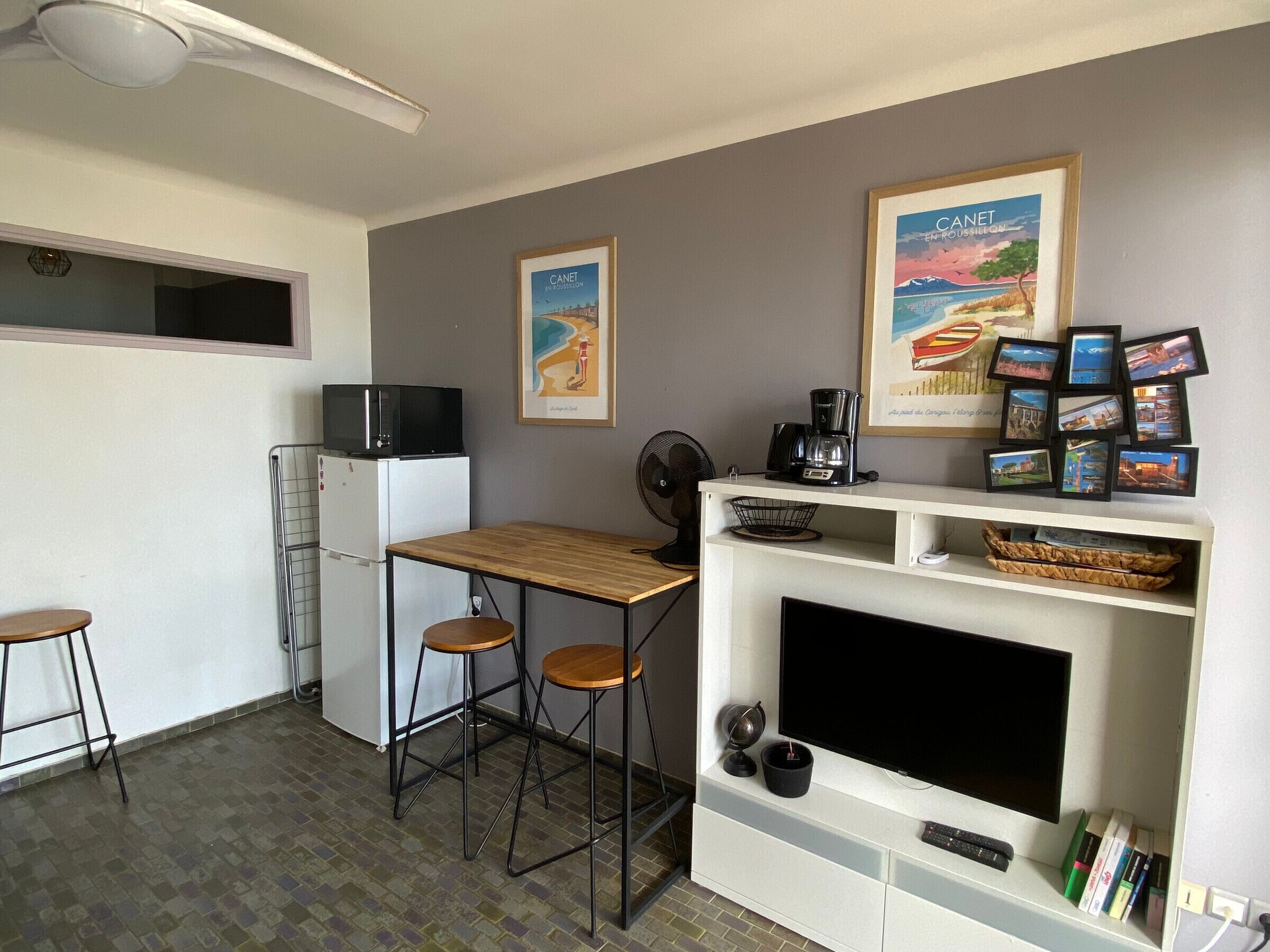 A compact kitchen with a microwave, refrigerator, and a small dining table with stools. There are framed pictures on the wall and a shelf with books and a coffee maker.