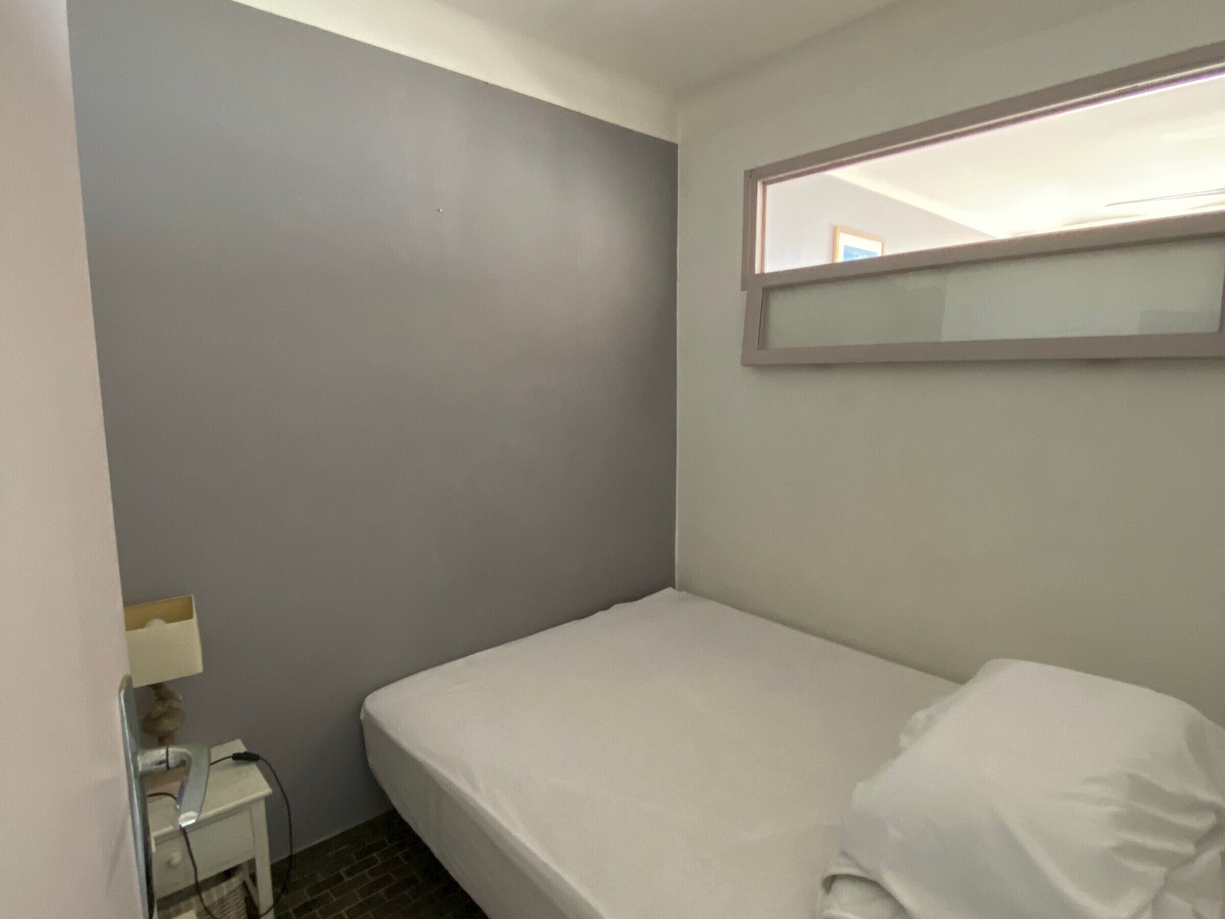 A small, single-bed room with a grey wall, a bedside table with a lamp, and a window with a mirror above the bed.