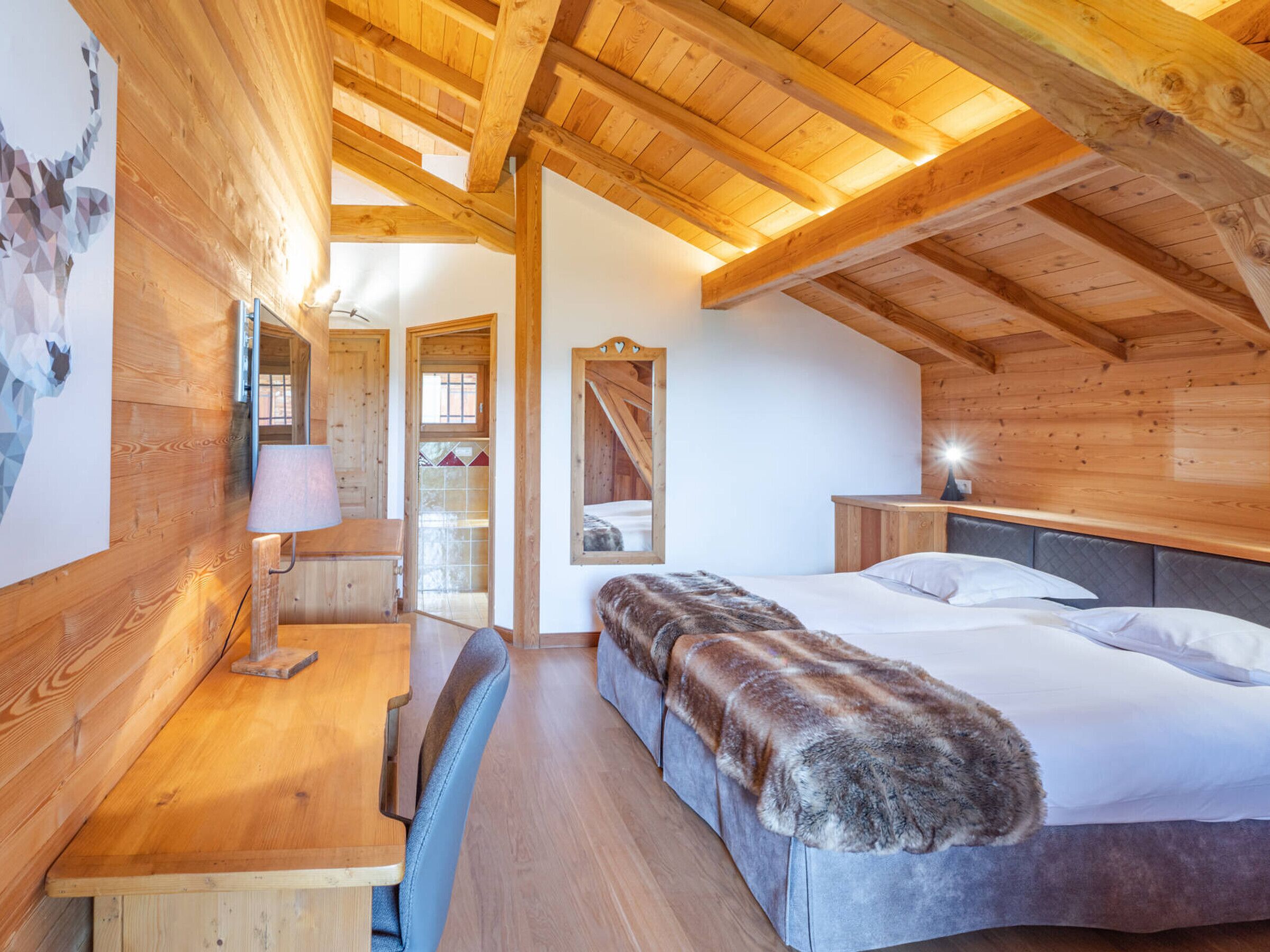 A cozy room with a wooden ceiling, a bed with a fur throw, a desk with a lamp, and a chair.