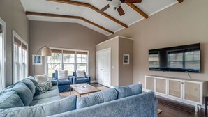 Un salon spacieux avec un grand canapé d’angle, une table basse et un ventilateur de plafond. Un téléviseur est fixé au mur et une fenêtre laisse entrer la lumière naturelle.