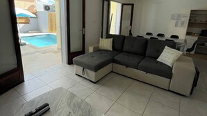 Un salon moderne avec un canapé d’angle, une table basse en marbre et une vue sur la piscine, visible à travers la porte vitrée.