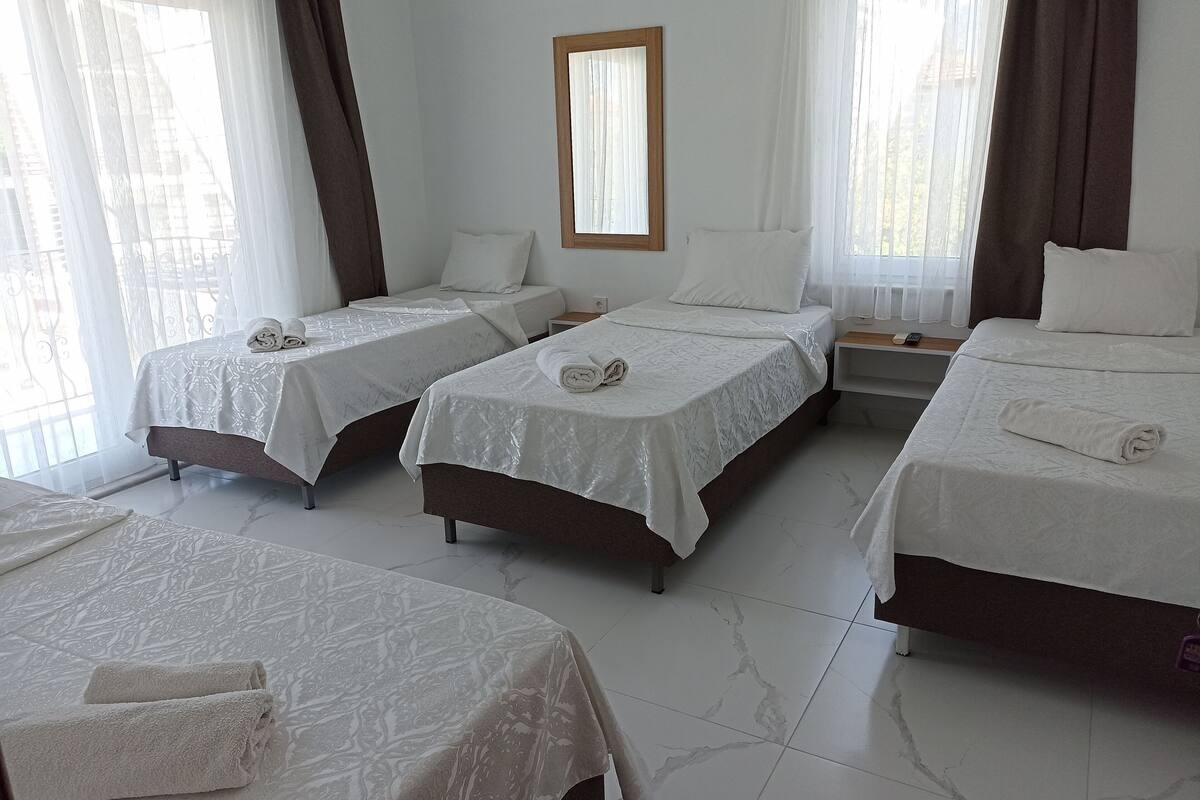 A hotel room with three single beds, each with a white bedspread and rolled towels. There is a wooden-framed mirror on the wall, and the room has white curtains.