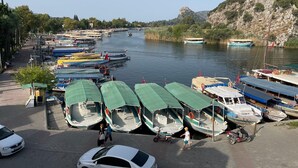 A marina with several boats docked, a paved walkway, and a hilly landscape in the background.