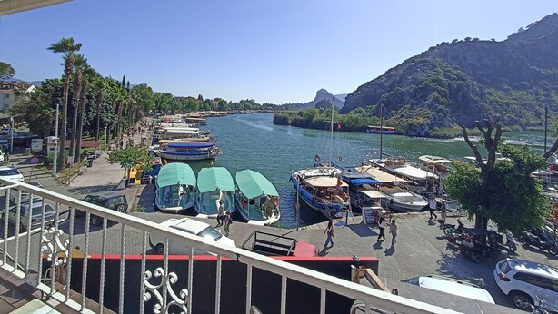 A riverside view with boats docked, a pedestrian walkway, and a mountainous backdrop.