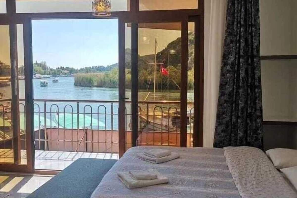 A hotel room with a bed, a view of a river, and a balcony with a railing.