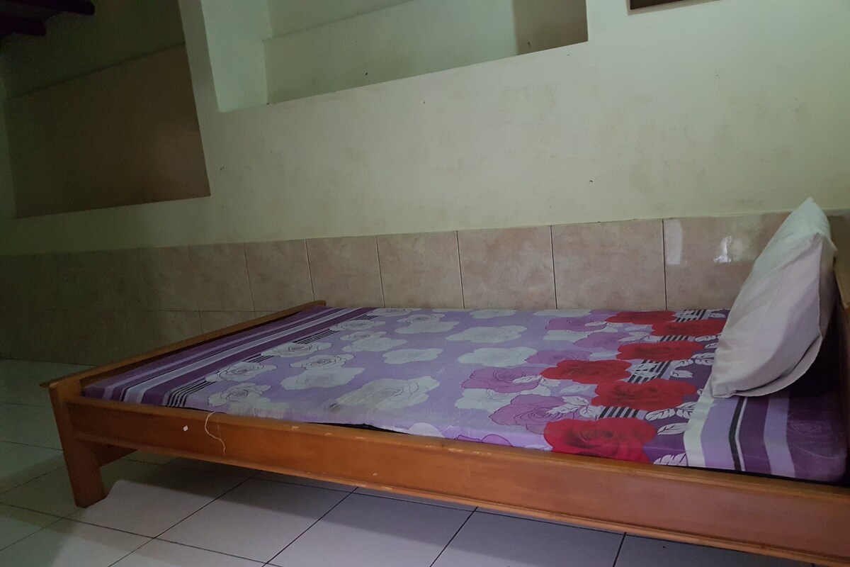 A single bed with a floral-patterned bedspread and a pillow, in a room with tiled flooring and a plain wall.