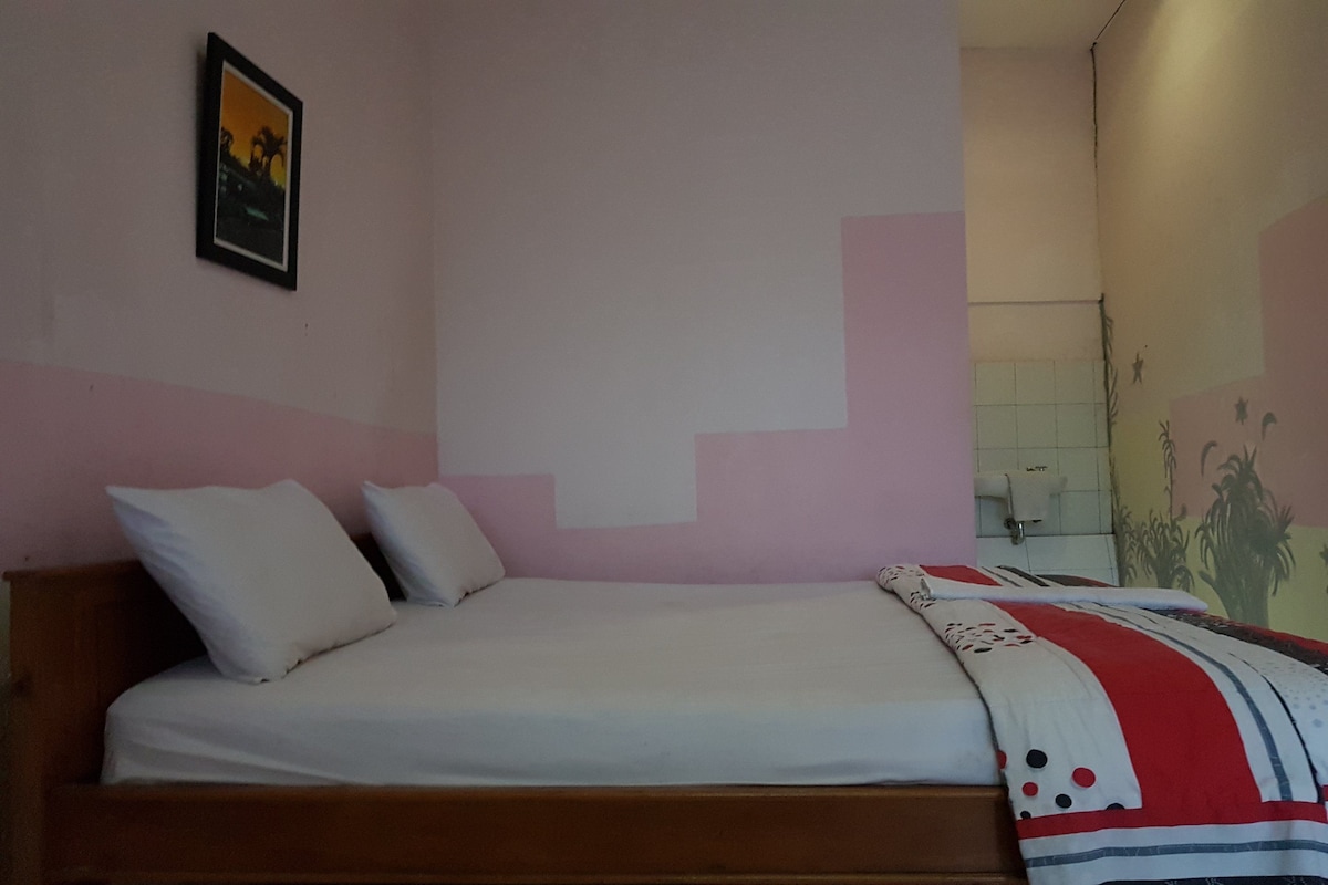 A bedroom with a bed, pillows, a blanket, a framed picture on the wall, and a bathroom with a towel dispenser.