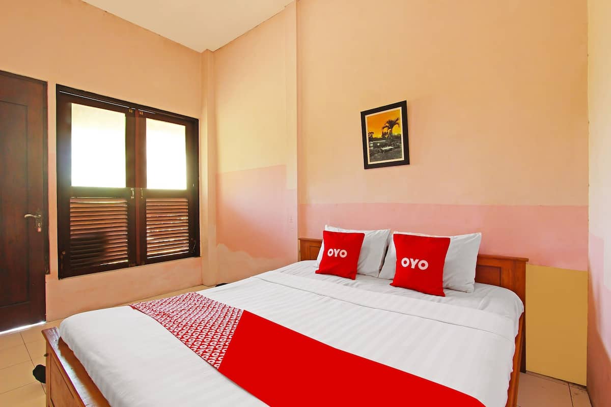 Un letto rifatto con copriletto rosso e bianco, due cuscini con la scritta "OYO", un quadro incorniciato appeso alla parete e una finestra con persiane.
