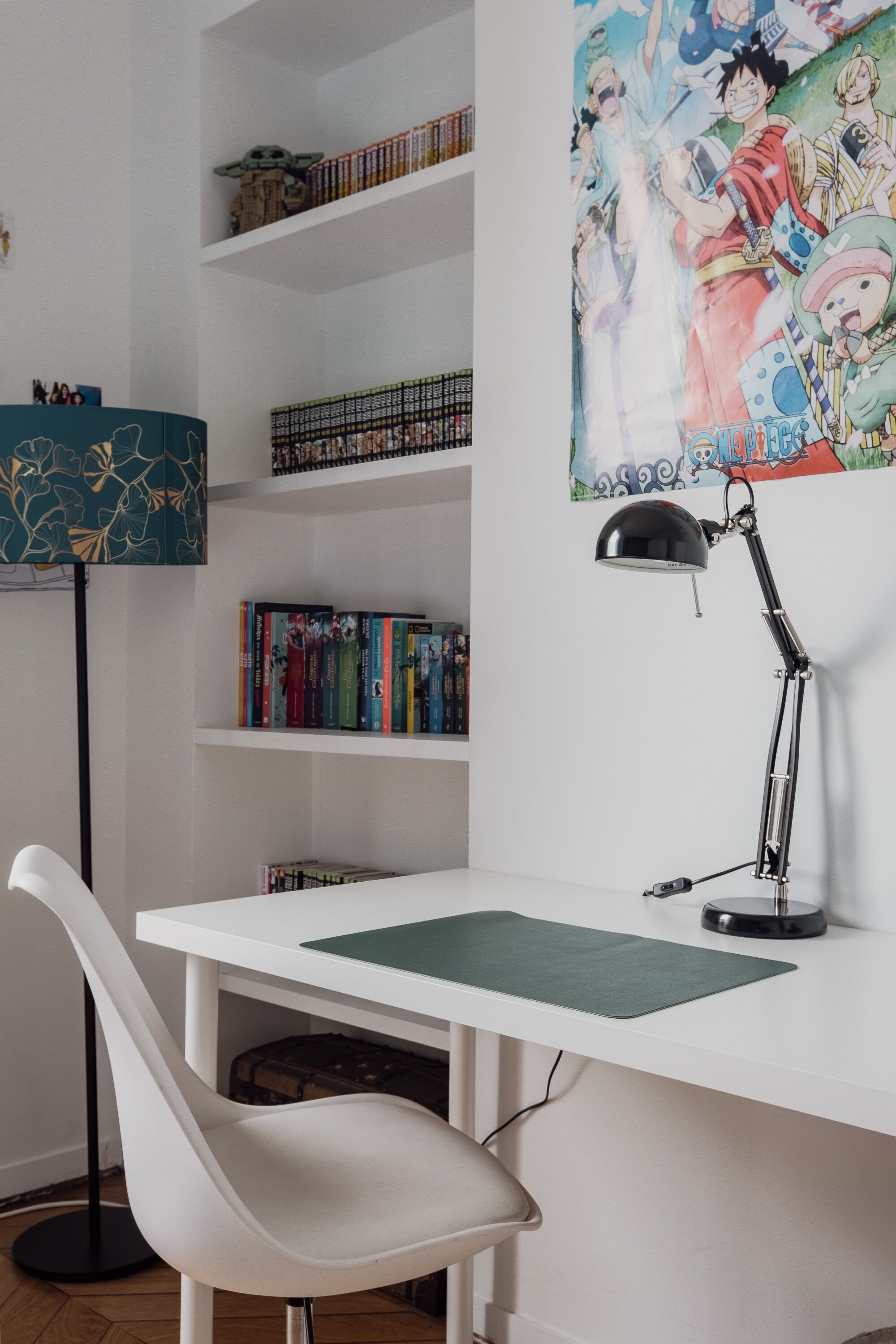 Un espace de travail avec un bureau blanc, une lampe de bureau noire et une chaise blanche. On peut voir des étagères remplies de livres et une affiche d’anime encadrée.
