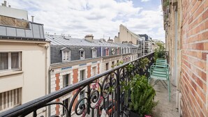 Une rue étroite en milieu urbain, bordée de maisons à étages, avec un garde-corps de balcon en métal et une chaise verte.