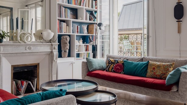 Un salon chaleureux avec une cheminée, une bibliothèque, un canapé agrémenté de coussins colorés et une grande fenêtre donnant sur l’extérieur.