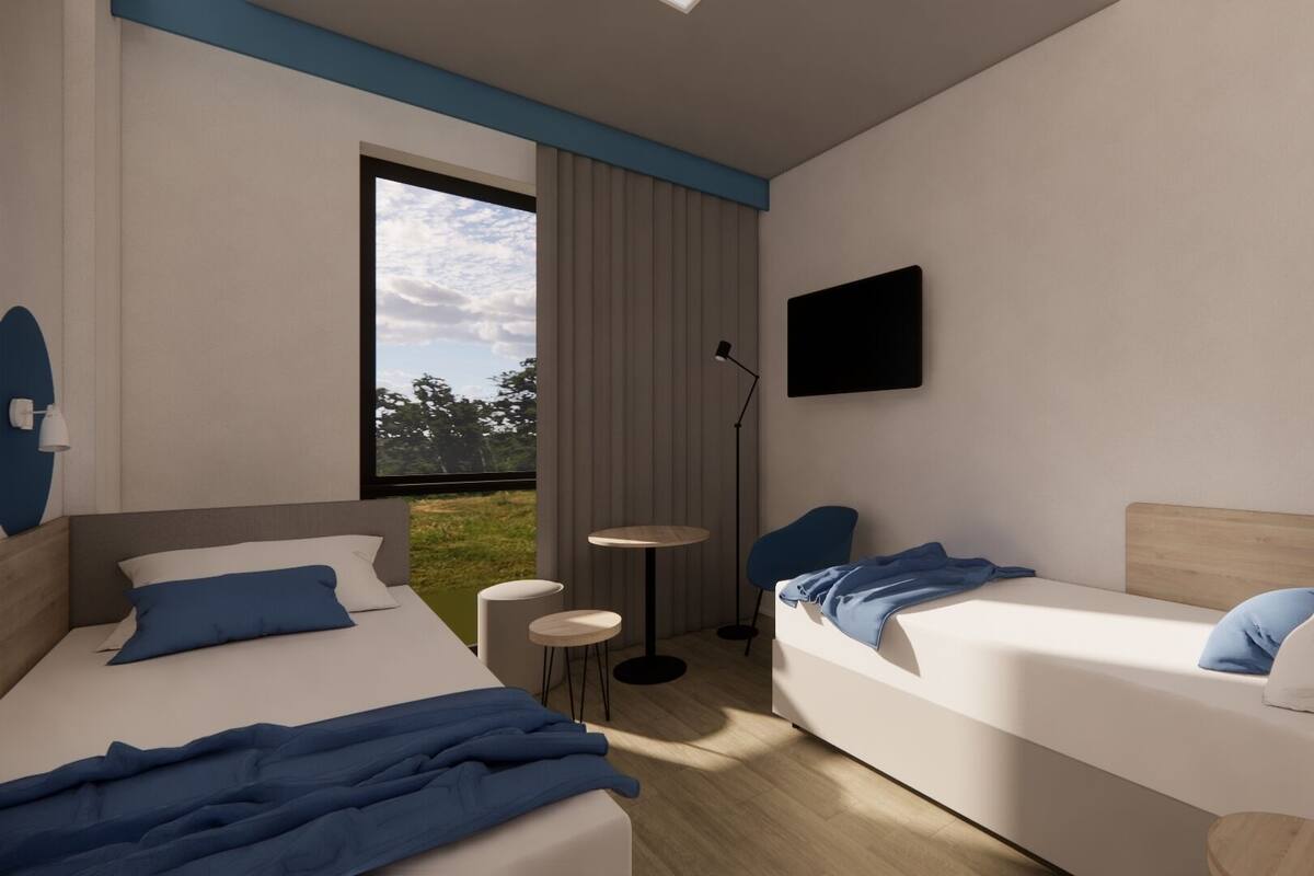 A modern hotel room with two beds, a TV, a small table, and a window with a view of trees.