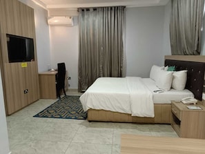 A hotel room with a bed, a desk, a chair, a TV mounted on the wall, and a window with curtains. - Eleventh House Hotel and Suites (Akure)