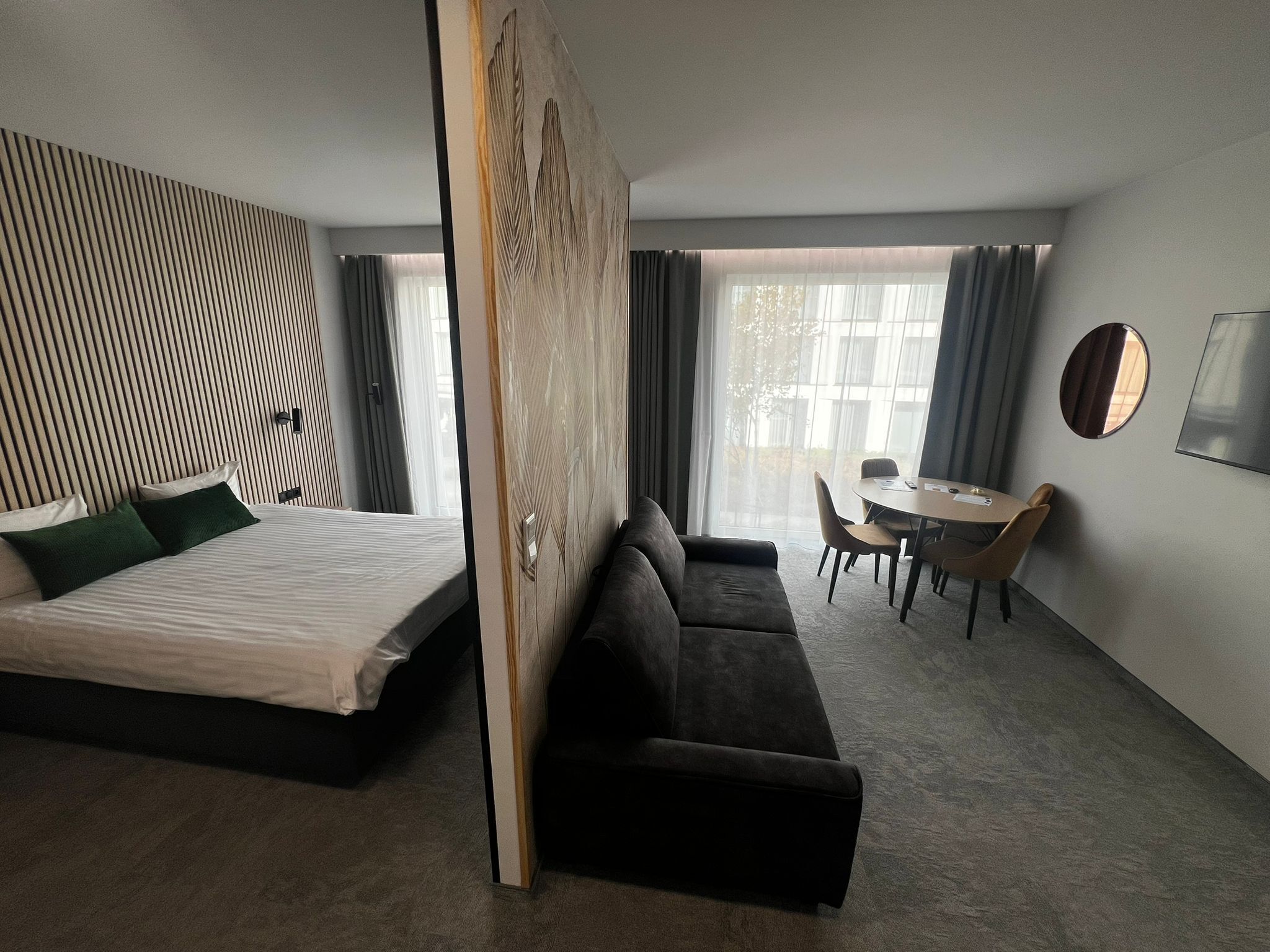 Una camera d'hotel moderna con un letto grande, un divano, un'area pranzo con tavolo e sedie e un'ampia finestra con tende.