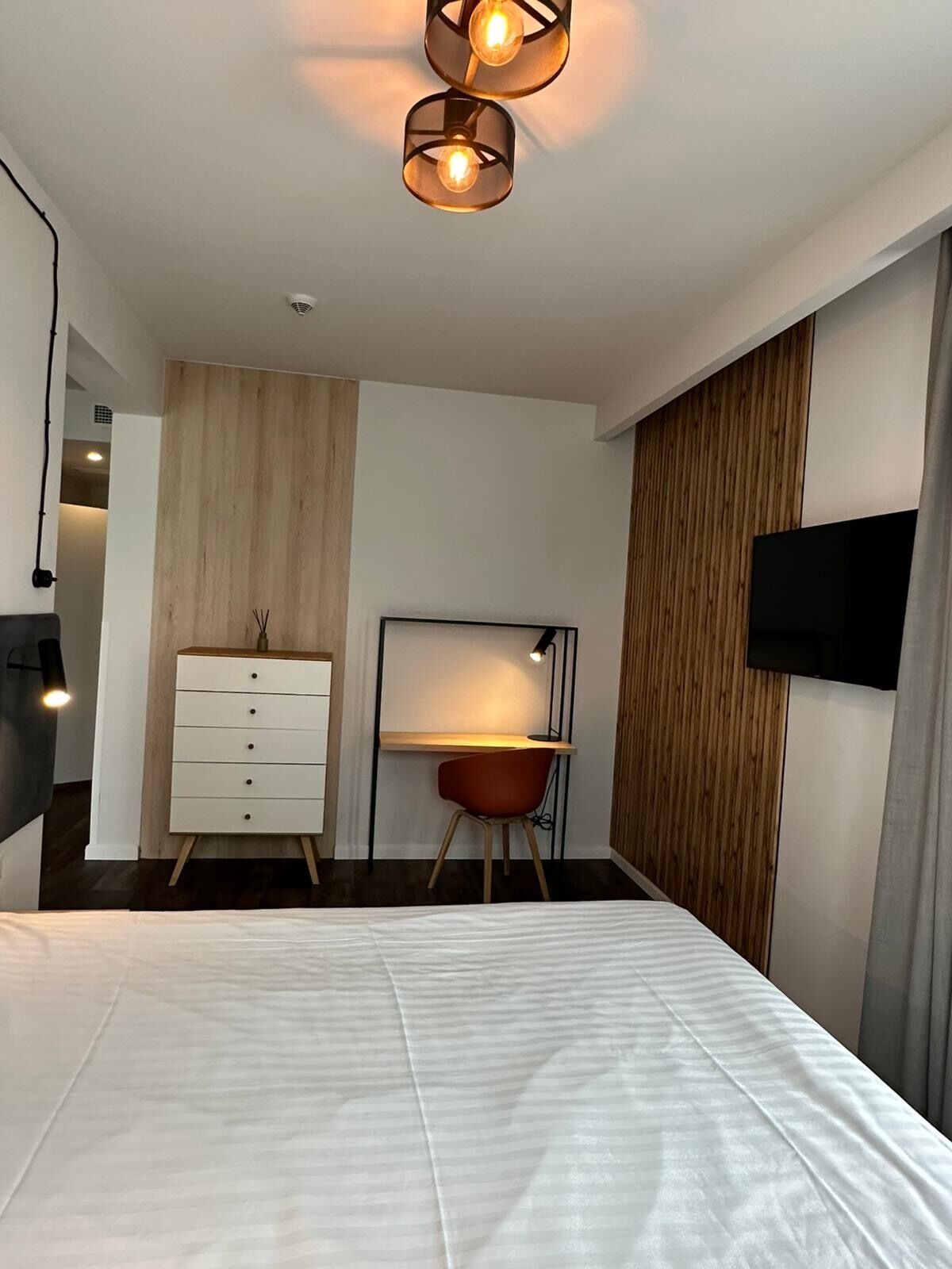 A modern bedroom with a bed, a desk, a chair, a dresser, and a TV mounted on the wall.
