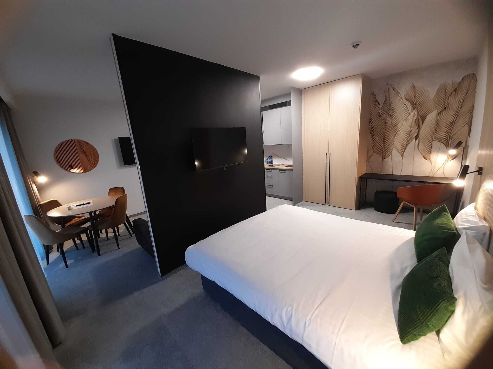 A modern hotel room with a large bed, a desk with chairs, a TV, and a kitchenette.