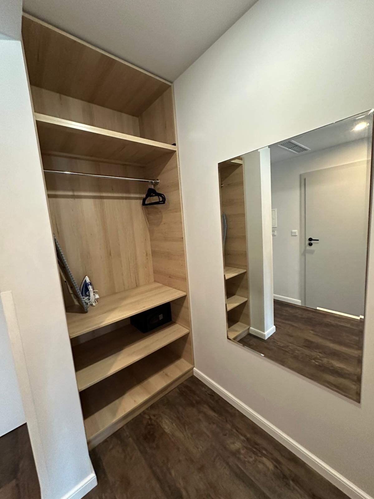 A walk-in closet with wooden shelves and a hanging rod, a small ironing board, and a mirror on the wall.