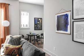 A living room with a grey sofa, a coffee table, and a dining table with chairs. There are framed artworks on the walls and a window with blinds.