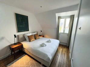 A bedroom with a bed, a nightstand, a lamp, a window with curtains, and a radiator. - Copernic Line | Elegant 3BR Apartment in Paris (Paris)