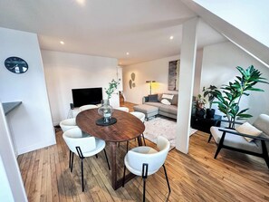 A modern living room with a wooden dining table, white chairs, a sofa, and a TV. - Copernic Line | Elegant 3BR Apartment in Paris (Paris)