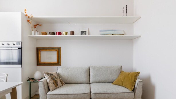 Ett modernt vardagsrum med en grå soffa, ett litet bord med en lampa och en köksdel med en spis och hyllor ovanför.
