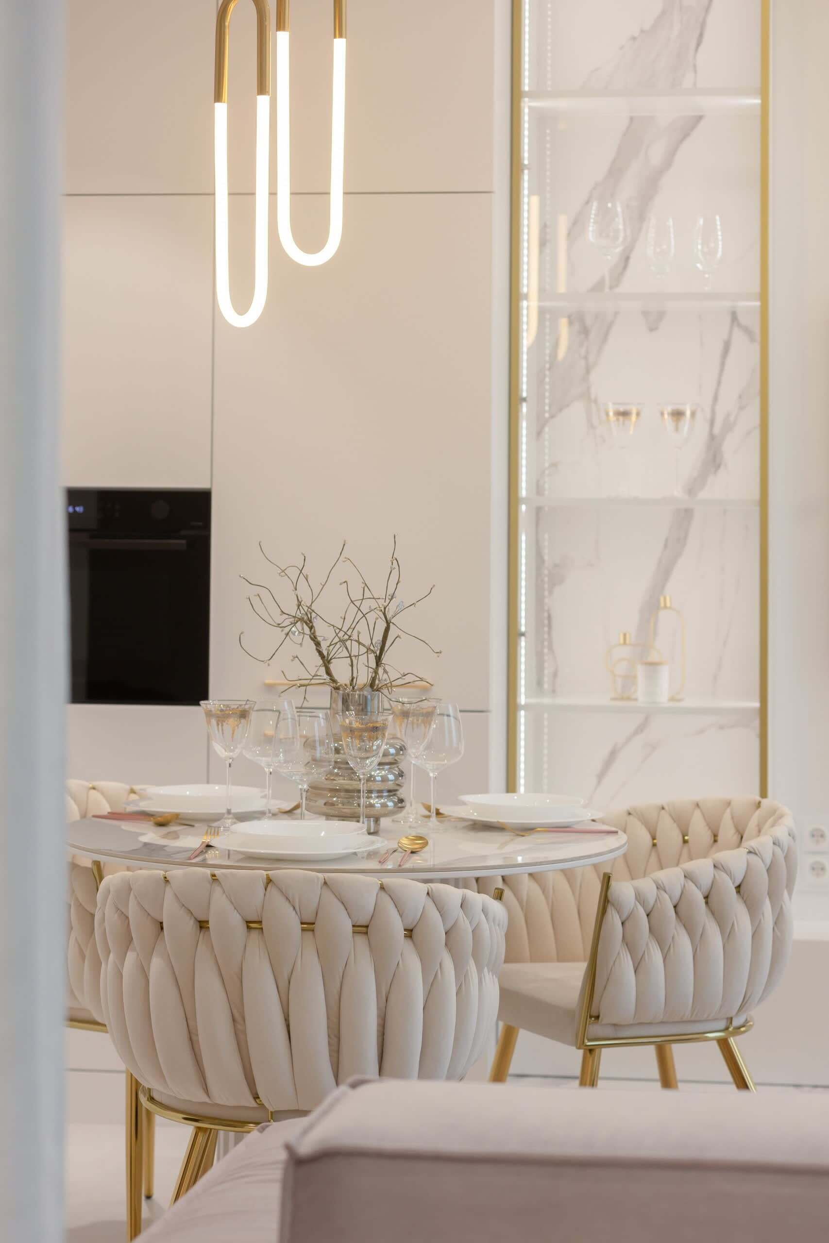 A modern dining area with a round table, white upholstered chairs, and a glass vase with branches.