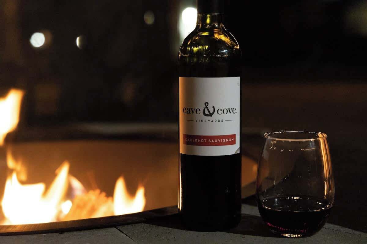 A bottle of Cave & Cove Vineyard Cabernet Sauvignon and an empty glass next to a fire.
