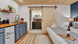 A modern hotel room with a wooden ceiling, a kitchen area with blue cabinets, a bed with a wooden headboard, and a bedside table with a bottle and glasses.