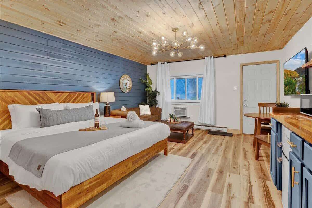 A bedroom with a wooden bed, a nightstand, a bedside table, a clock, a window with curtains, a TV, and a kitchen area with cabinets and a countertop.