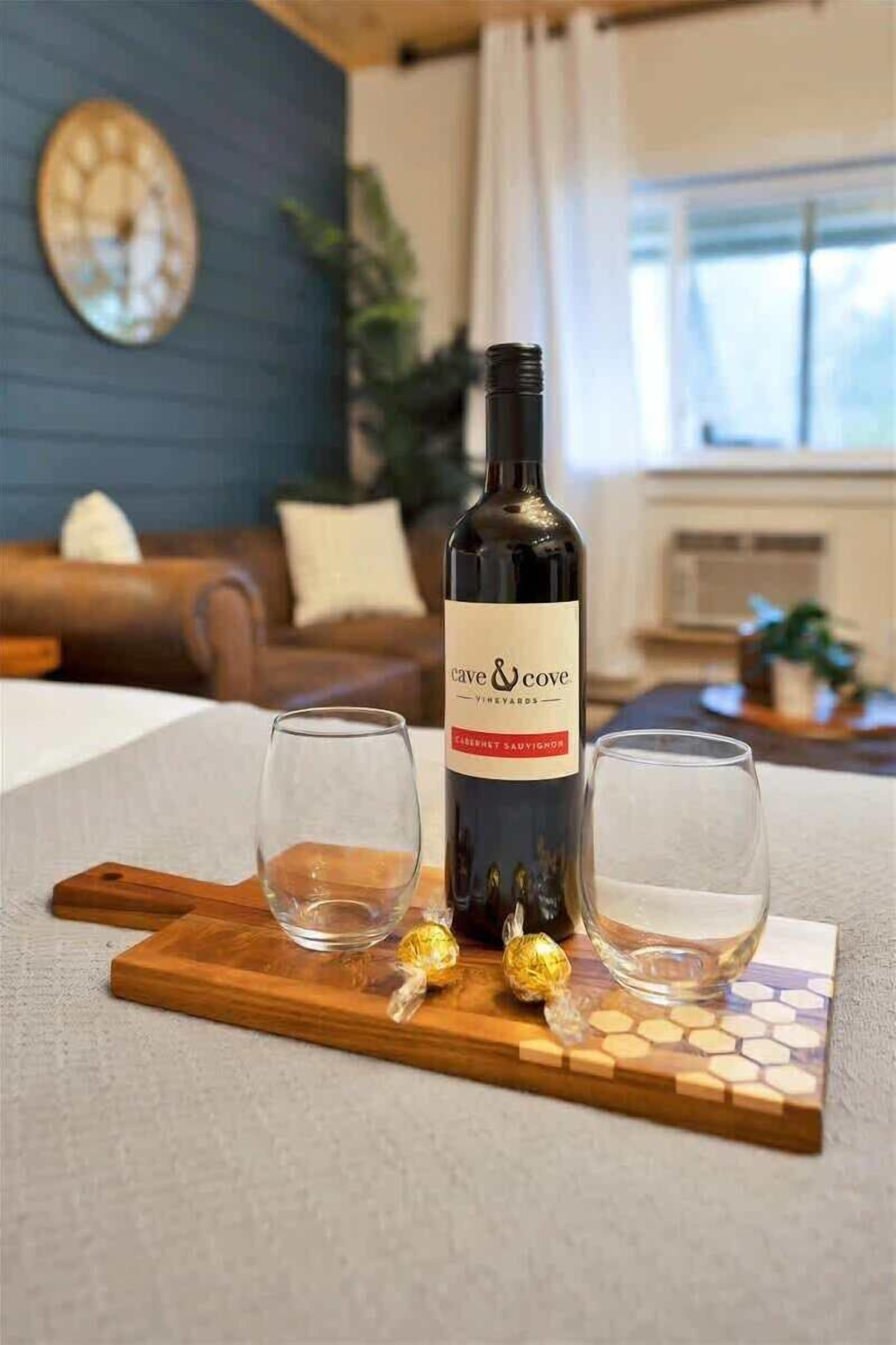 A bottle of Cave & Cove Vineyard Cabernet Sauvignon on a wooden board with two glasses, in a room with a blue wall, a clock, and a couch.