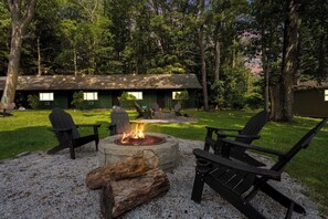 Un chalet doté d’un foyer entouré de fauteuils Adirondack, situé dans une zone boisée, avec un bâtiment écologique en arrière-plan.