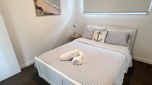 Una cama bien hecha con ropa de cama a rayas, una toalla enrollada, una mesita de noche con lámpara y un cuadro en la pared.