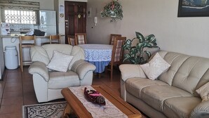 Una sala de estar con sofá, una mesa de comedor con sillas, un área de cocina con refrigerador y un ventanal grande.