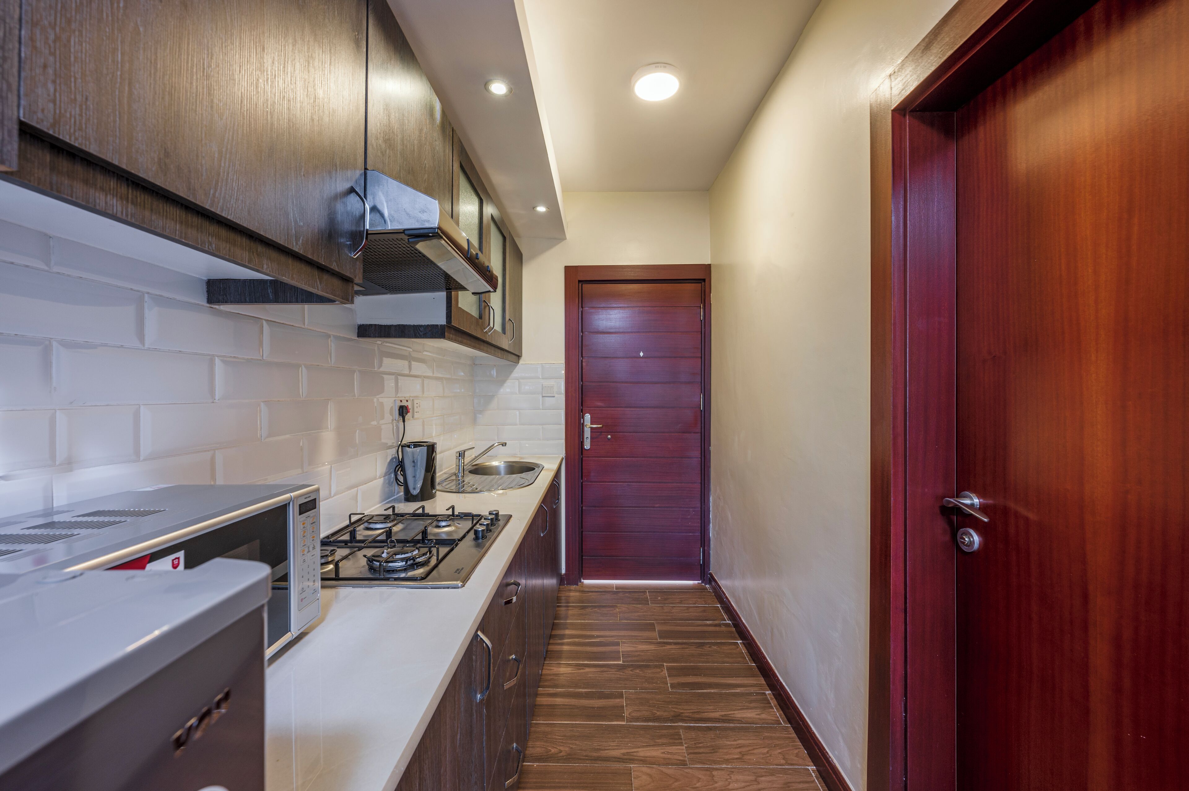 A narrow kitchen with a gas stove, a microwave, and a sink. There is a wooden door at the end of the hallway.