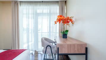 Une chambre d’hôtel avec un lit, un bureau et une chaise, une plante en pot et une grande fenêtre avec des rideaux.