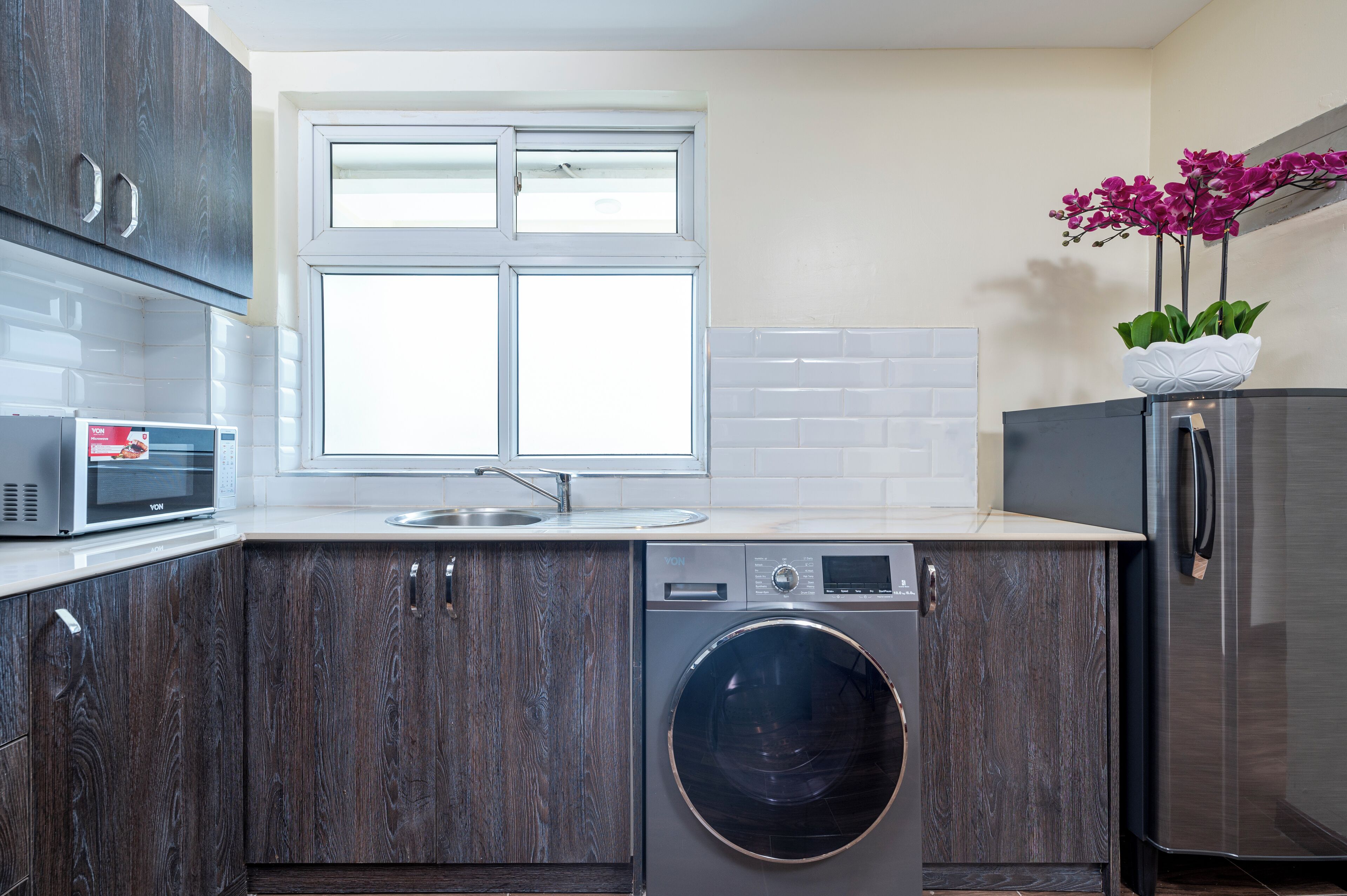 Una cocina moderna con armarios de madera oscura, un microondas integrado, un refrigerador de acero inoxidable y una lavadora.
