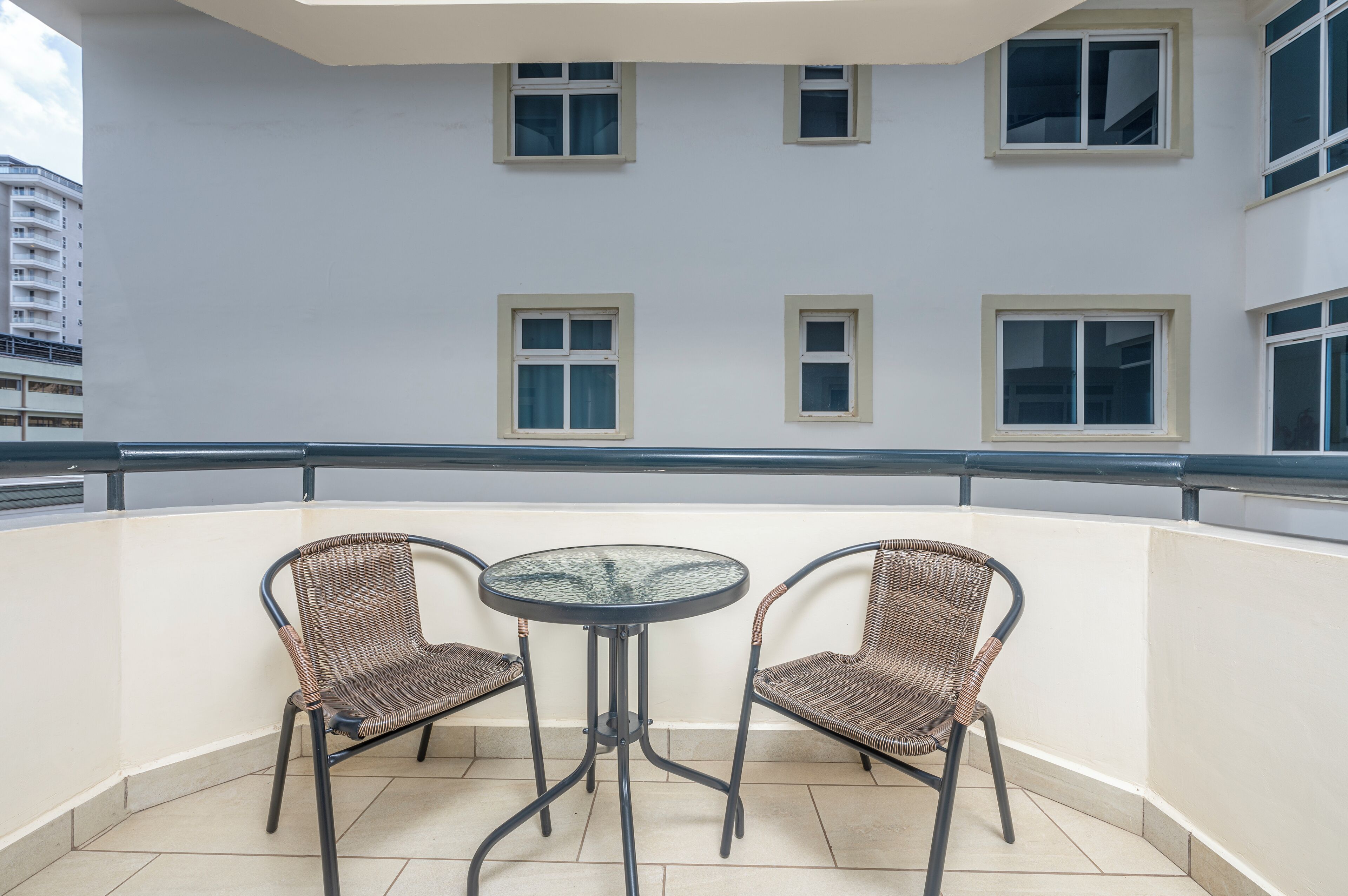 A balcony with two wicker chairs and a glass table, white walls, and multiple windows.