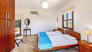 A bedroom with two beds, a wooden wardrobe, a small table, a mirror, and a window with a view. - Villa Anthia (Pegeia)