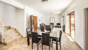 A spacious living area with a dining table and chairs, a sofa, and a TV. There is a staircase on the left and a door leading to another room. - Villa Anthia (Pegeia)