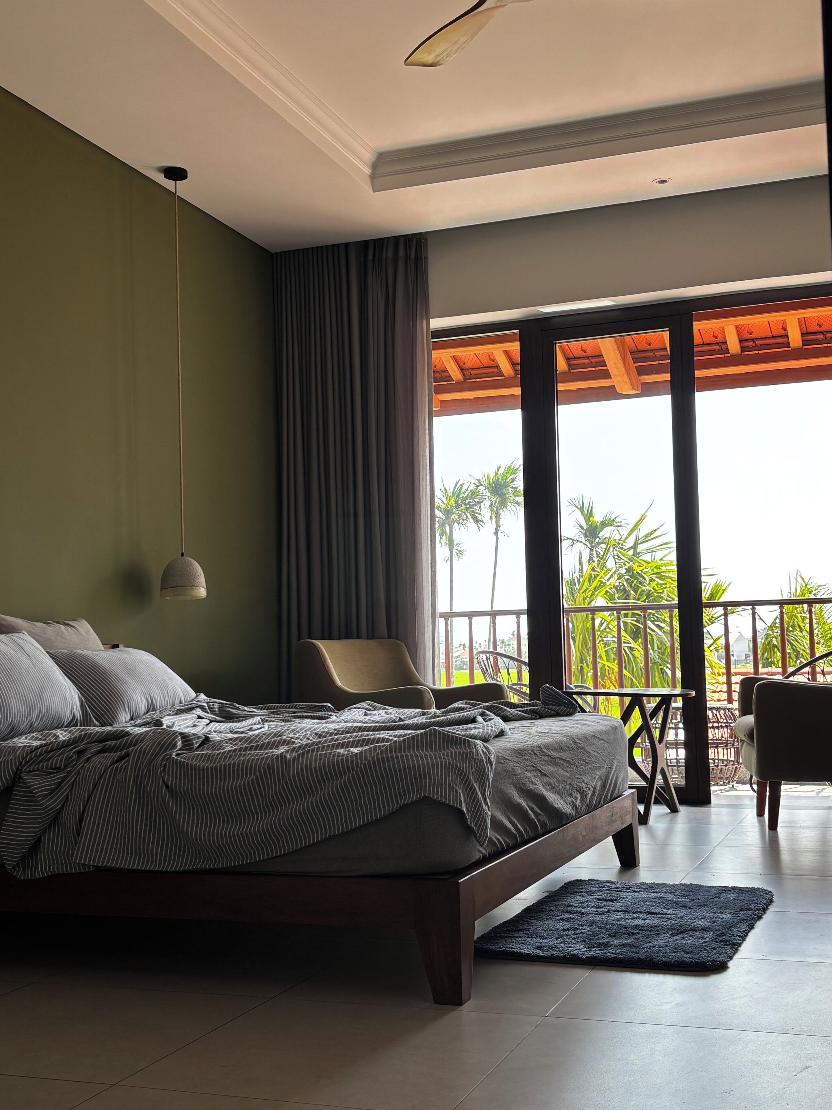 Een slaapkamer met een groot bed, een stoel, een balkon met uitzicht op palmbomen en een plafondventilator.