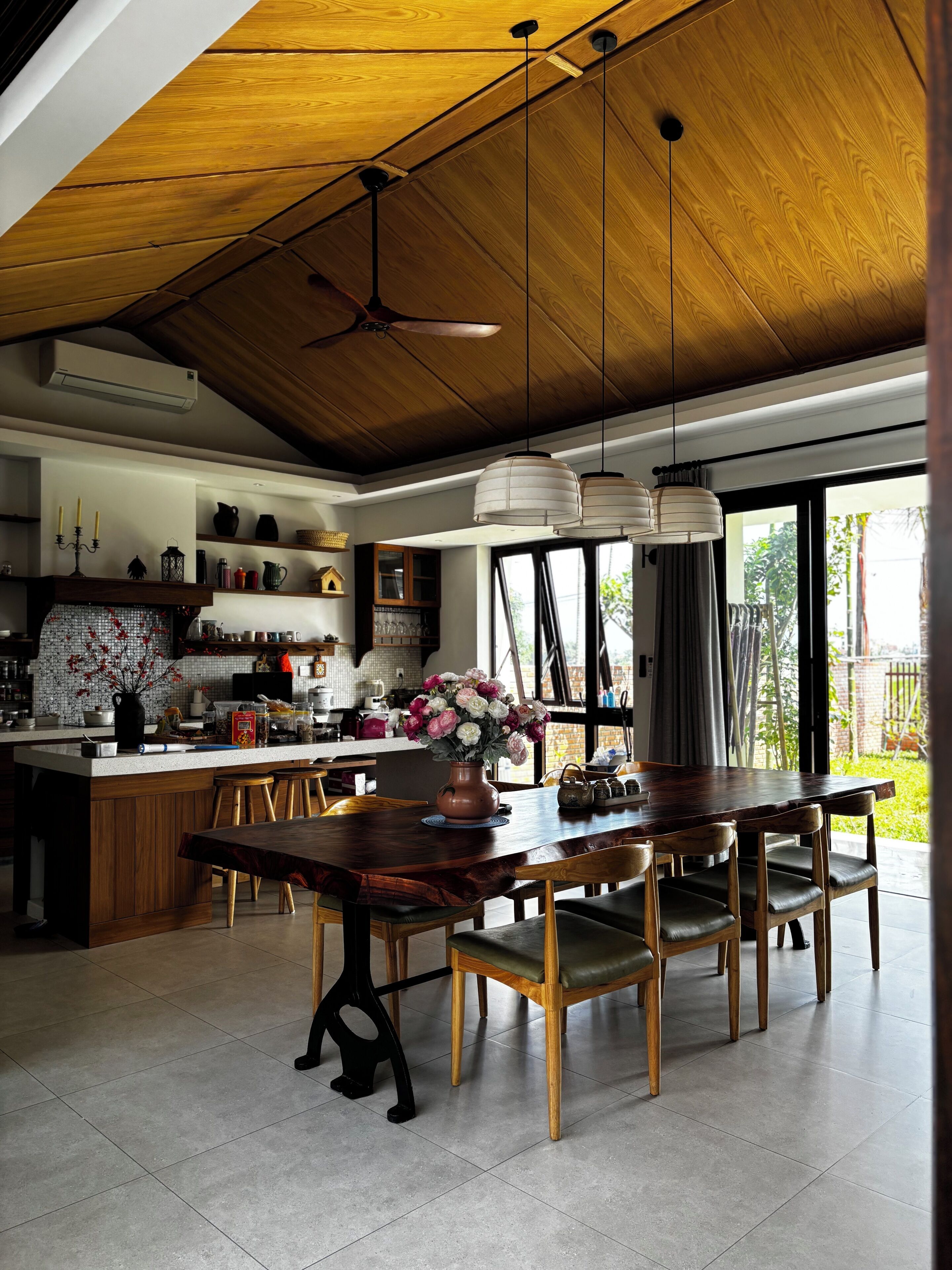 Een ruime keuken met een houten plafond, een grote eettafel met stoelen en uitzicht op het buitengebied door grote ramen.