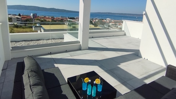 A rooftop terrace with a glass balustrade, a black coffee table with drinks, and a view of the sea and distant buildings. - Villa With Panoramic Views on the Sea, Islands, City and Hill (Kaštel Sućurac)