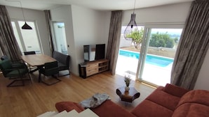 A modern living room with a wooden dining table, green chairs, a brown sofa, a TV, and a view of a swimming pool through large windows. - Villa With Panoramic Views on the Sea, Islands, City and Hill (Kaštel Sućurac)