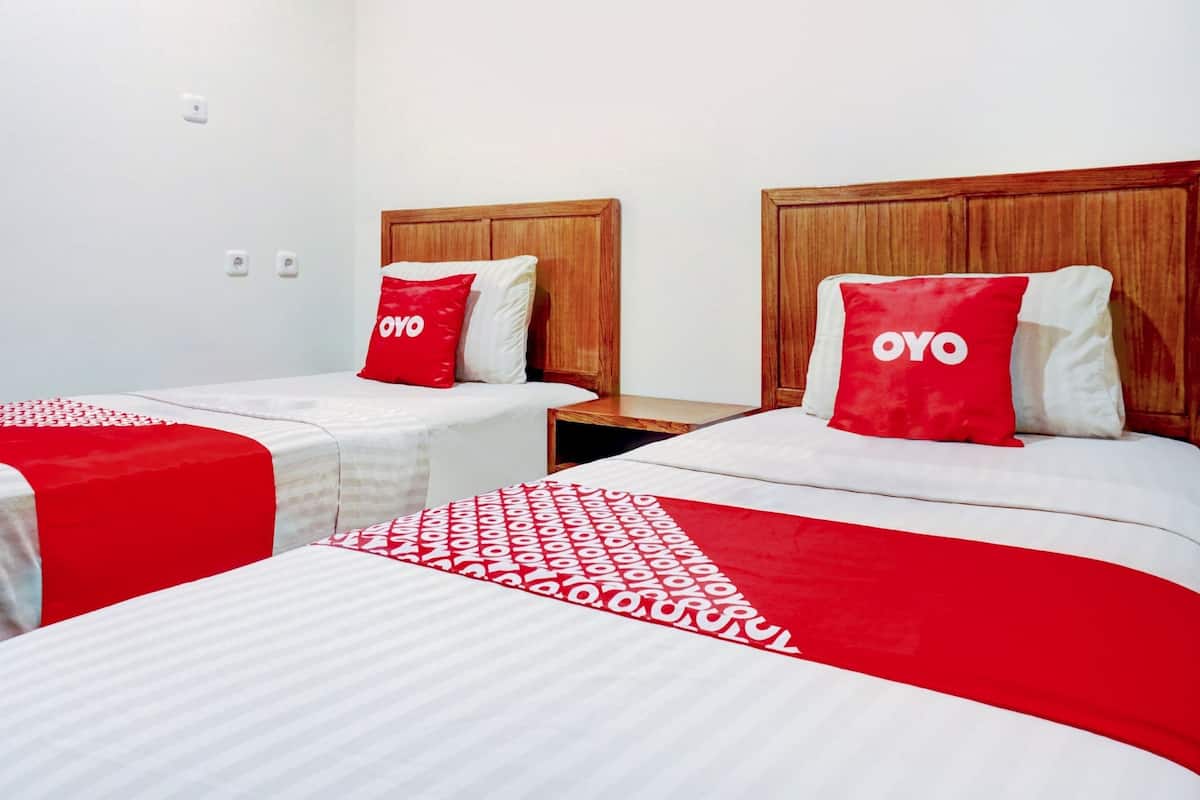 Habitación de hotel con dos camas, cada una con cabecero de madera, ropa blanca y almohadas rojas con la palabra "OYO".