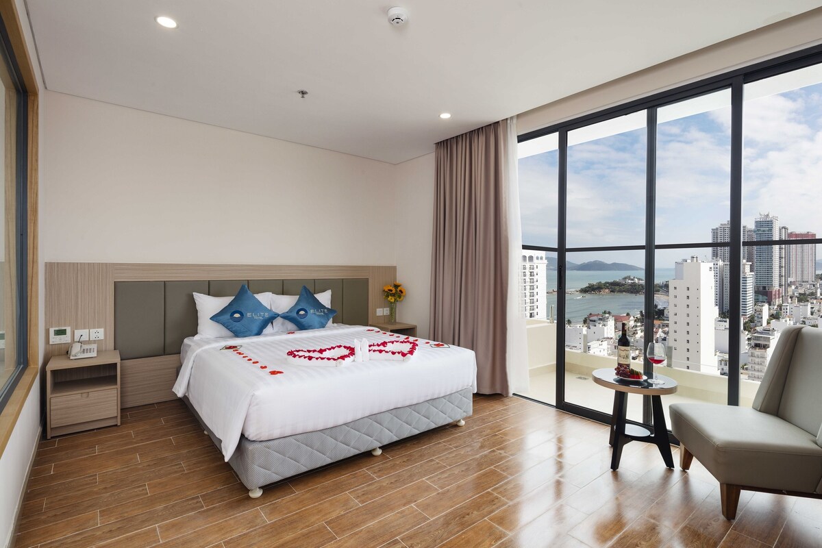 Camera d'albergo con un letto grande, un comodino, un tavolino con una bottiglia e un bicchiere, una sedia e un'ampia finestra vista città.
