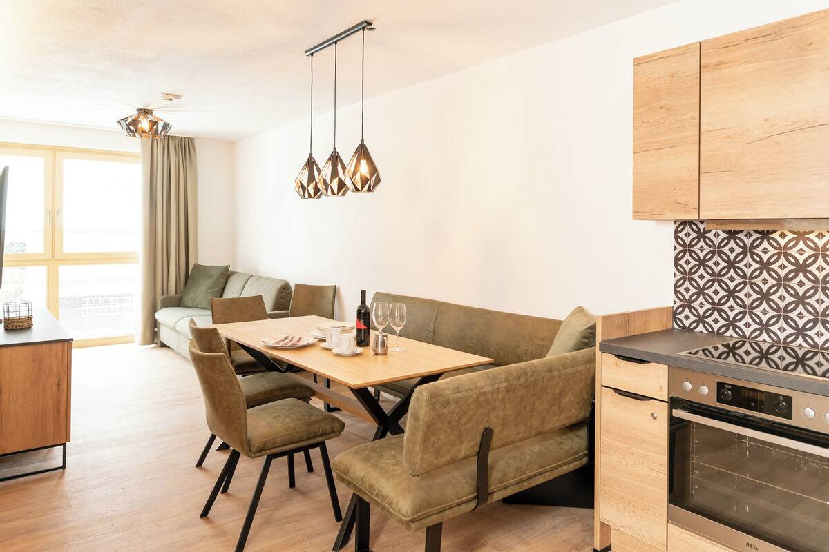 A modern kitchen with a built-in oven, a dining area with a wooden table and cushioned chairs, and a sofa in the background.
