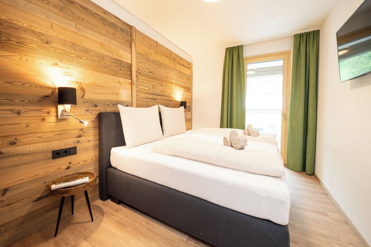A modern hotel room with a wooden wall panel, a bed with white linens, a small stool, and a flat-screen TV mounted on the wall.