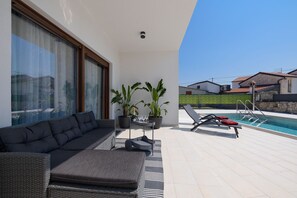 Un espacio al aire libre moderno con un sofá, una mesita con vasos, plantas en macetas y una piscina con sillas de descanso.